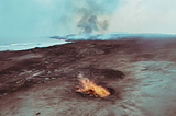 A small fire smoulders on a piece of arid land, with the sea in the distance, and smoke and ash floating in the air.