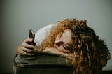 A woman with long curly red hair is slumped over a cushion, looking at her mobile phone. She seems sad or melancholy as she stares at her phone.