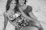 Black and white photo of the author, a young woman, sitting across her mothers lap on a sandy beach.