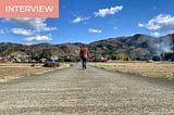 A man is turning his back at the camera, walking in the middle of a deserted road in the Japanese countryside, towards the mountains. The sky is blue.