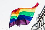 Pride flag waving in front of a city building. Image is black and white, except for the rainbow colors of the Pride flag.