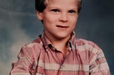 Author at age 6 with short blondish-brown hair. School photo, smiling and wearing a light red button up.
