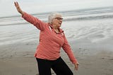 An older woman on the beach, smiling, with her arms in a position that depicts the freedom and joy she feels at the shoreline