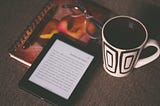 Photo of an e-reader, notebook and coffee cup on wooden surface