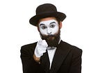 Photo of a mime artist/jester pointing a white-gloved finger at the camera. He has a black beard and wears a hat.