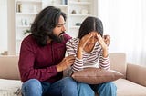 A man and a woman are sitting on a sofa together. The woman has her head in her hands, clearly upset. The man has his arms around her, comforting her.