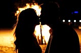 A sexy teenage girl and guy share a secret romantic kiss silhouetted in front of a bonfire on a riverside beach on a hot Summer night.