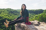 Lisa Le sits on a rocky bluff overlooking a lush forest.