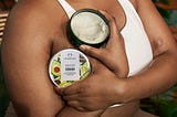 A dark-skinned individual wearing a white tank top shows the lid and inside of the tub of The Body Shop’s Avocado Body Butter.