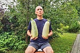 The author, József Manhertz, sits on a chair in his garden in summer in green shorts and a dark blue T-shirt. Lush green bushes and trees surround it. He is holding two huge zucchini in both hands. Meanwhile, he looks at the camera with his head held high, with a funny forced proud expression.