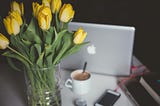 View of a desk with a computer, coffee, a charging phone and beautiful yellow tulips.