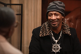 Comedian Katt Williams, wearing a black and grey beanie, a black shirt jacket with a patterned butterfly collar, and a large medallion in the shape of a captain’s wheel, smiling during a podcast taping.
