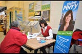 Former MP Claire Perry conducting a constituency surgery meeting with a constituent in a public building with a poster to the left which says “Claire Perry (headshot) How can I help?”