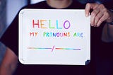 A light-skinned human being in a black shirt holds up a sign with rainbow text reading “HELLO MY PRONOUNS ARE”