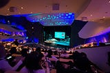 There is a stage in the center of the image and sitting stands around it. There is a screen on stage and people sitting on the the stands.