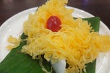 On a white plate, over a banana leaf yellow threads are placed on a custard-texture pudding with a cherry on the top.