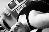 Black-and-white selfie of Alex lying on an exam table, a pair of gauze pads on their forehead with a small dot of blood at the centre of each. Behind Alex’s head, the metal arm of some kind of medical imaging tool arcs upward, toward a pair of bulky cathode ray tube monitors which are giving off that weird, distinctly clinical vibe of outdated futurism.