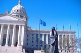 Bronze sculpture of a Native American woman in front of the neoclassical facade of the Oklahoma State Capitol building.