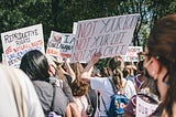 Image of women at a protest, holding signs supporting women’s reproductive rights. Masked protestors hold pro-choice signs and are viewed from the back, during daytime amid green trees.