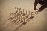 CORE VALUE wood word on compressed or corkboard with human’s finger at S letter