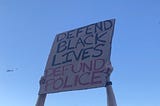 Hands thrust a protest sign reading “Defend Black Lives Defund Police” into the sky, as an LAPD helicopter flies overhead.