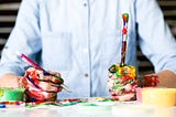 Photo of messy hands covered in paint, holding a pencil and paintbrush