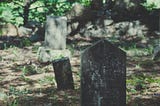 Picture of several very weathered gravestones in a cemetery in the woods.