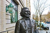 Bronze statue of a man from the nineteenth century. He’s looking forward, and stands next to a decorative copper lamp.