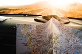 A map rests against a car dashboard as a sunset is visible through the windshield. Photo by Julentto Photography on Unsplash