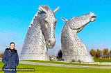 A brunette in a blue coat standing in front of two large metallic horse heads and smiling at camera on a sunny day.