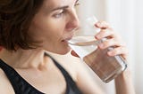 A young woman with short brown hair and wearing a black tank top is drinking a glass of water.