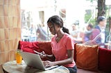 A young woman is sitting inside a restaurant in front of her laptop, while looking at her cellphone.