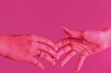 Two hands reaching for one another, seen under pink light against a bright pink background