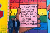 An older Black woman standing next to an Asian college student in front of a mural on a brick wall. The mural is in bright colors and shows a large hand holding a card with a quote.