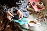 Woman scrolling through her cell phone with a cup of coffee and books next to her.