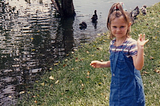 A young girl waves alongside the edge of a lake, with ducks in the distance.