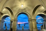 Photo of Trieste’s Piazza dell’Unita d’Italia from behind a tri-arched dome, at sunset with a pink & blue sky in the background.