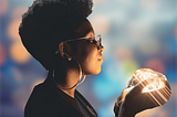 A profile of a Black woman wearing a black t-shirt, and large, thin silver hoop earrings has an afro. She is wearing thin silver colored eyeglasses and has eyes closed while holding a light in her hands near her face. The light from her hands; illuminates her face. Blurred background of multi-colored lights.