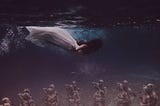 A woman in a white dress, diving close under the water surface, looking down, dark long hair is covering her face. On the ground are mannequins arranged as if they were a couple — but many of them.