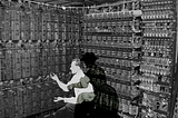 A black-and-white image depicting the early computer, Bombe Machine, during World War II. In the foreground, the shadow of a woman in vintage clothing is cast on a man changing the machine’s cable.