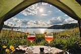 Image shows a view on a lake and mountains, and grass with wildflowers from inside a tent with and two cocktail glasses in the tent opening.