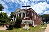 Historic red-brick First Presbyterian Church in Smithville, Texas, with a large cross standing in front beneath a bright blue sky filled with scattered clouds.