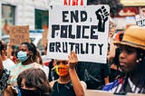 An image of a crowd of protesters. In the center, a woman with brown skin and a colorful face masks holds up a white sign with “End Police Brutality” in black writing and a Black Power fist.