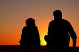 The silhouettes of an adult and a child sitting next to each other against a sunset