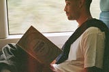 Loyle Carner sitting in a train seat where the window behind him shows the countryside passing by in a blur; Loyle is holding Yotam Ottolenghi’s cookbook “Jerusalem” in his hands and reading it