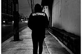 Man walking down the street, shoulders hunched, on his way to work. His jacket says “Maintenance” across the back.