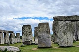 Ancient stone pillars and lintels at Stonehenge under a cloudy sky. The weathered stones stand on a grassy plain, with a narrow opening of blue sky visible between layers of grey cloud.