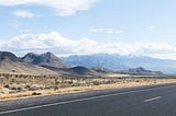 photo of set of mountains on the left with larger mountain the background. Foreground is a two lane highway in Utah