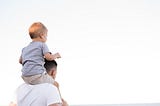 Man in white shirt carrying young boy on his shoulders, facing away from the camera, looking into the distance.