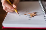 A person is writing in a notebook with a pencil. There are pencil shavings lying on the blank notebook page.